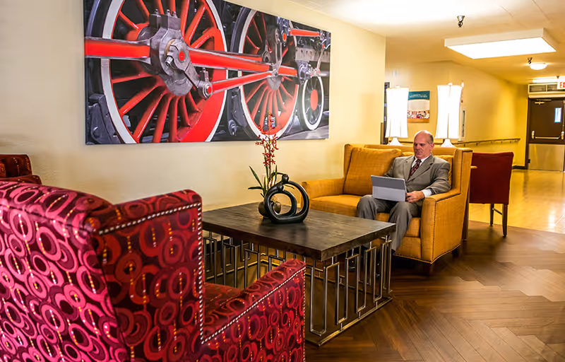 A man in a suit sitting on a mustard yellow armchair using a laptop in a hallway lounge area. The lounge features a red patterned armchair, a wooden coffee table with a decorative black sculpture and a plant, and a large wall art of red train wheels. The hallway has wooden flooring and is softly lit with wall-mounted lamps.