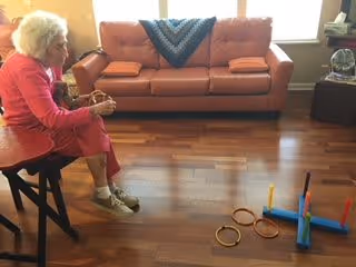 An elderly woman sitting on a chair in a living room with wooden flooring, playing a ring toss game. Behind her is a brown leather sofa with a blue knitted blanket draped over the backrest. A window is visible in the background letting in natural light.