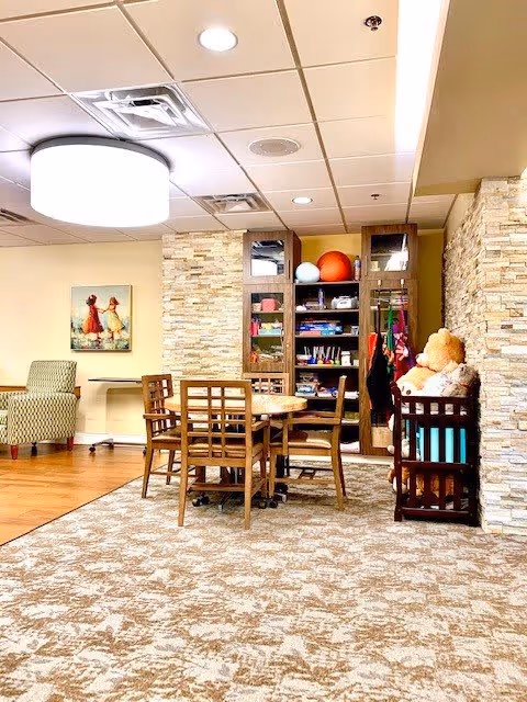 A cozy interior room with a round wooden table surrounded by six wooden chairs on a patterned carpet. Behind the table is a tall shelving unit filled with various items including books, games, and colorful balls. To the right, there is a wooden crib with a large teddy bear inside. The walls are decorated with light-colored stone accents and a framed picture of two children playing. The ceiling has recessed lighting and a large circular light fixture.