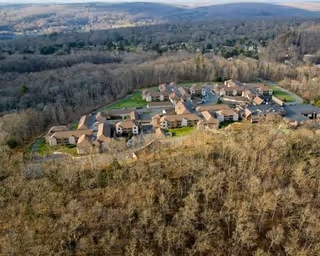 Aerial view of a senior living facility named The Watermark at East Hill, surrounded by dense forest and hills in the background. The facility consists of multiple connected buildings with brown roofs and beige walls, situated in a clearing with paved roads and parking areas.