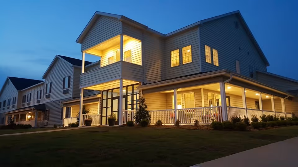 Exterior view of a two-story senior living facility building at dusk with lights glowing warmly from the windows and porch area. The building has white siding, a covered porch with white railings, and a balcony on the second floor. The surrounding area includes a grassy lawn and a paved walkway.