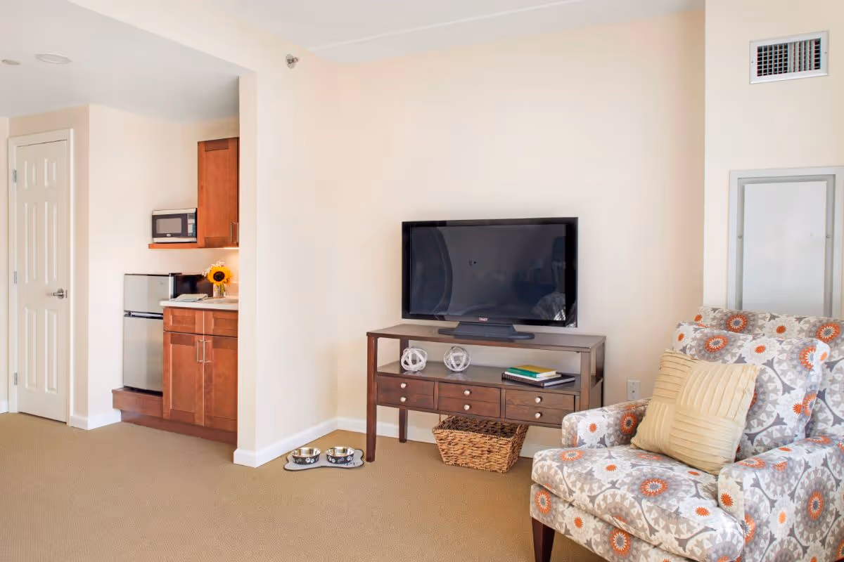 A cozy living area with a patterned armchair featuring a beige pillow, a wooden TV stand with a flat-screen television, decorative items, and books. To the left, there is a small kitchenette with a mini fridge, microwave, and wooden cabinets. The room has beige walls and carpeted flooring.
