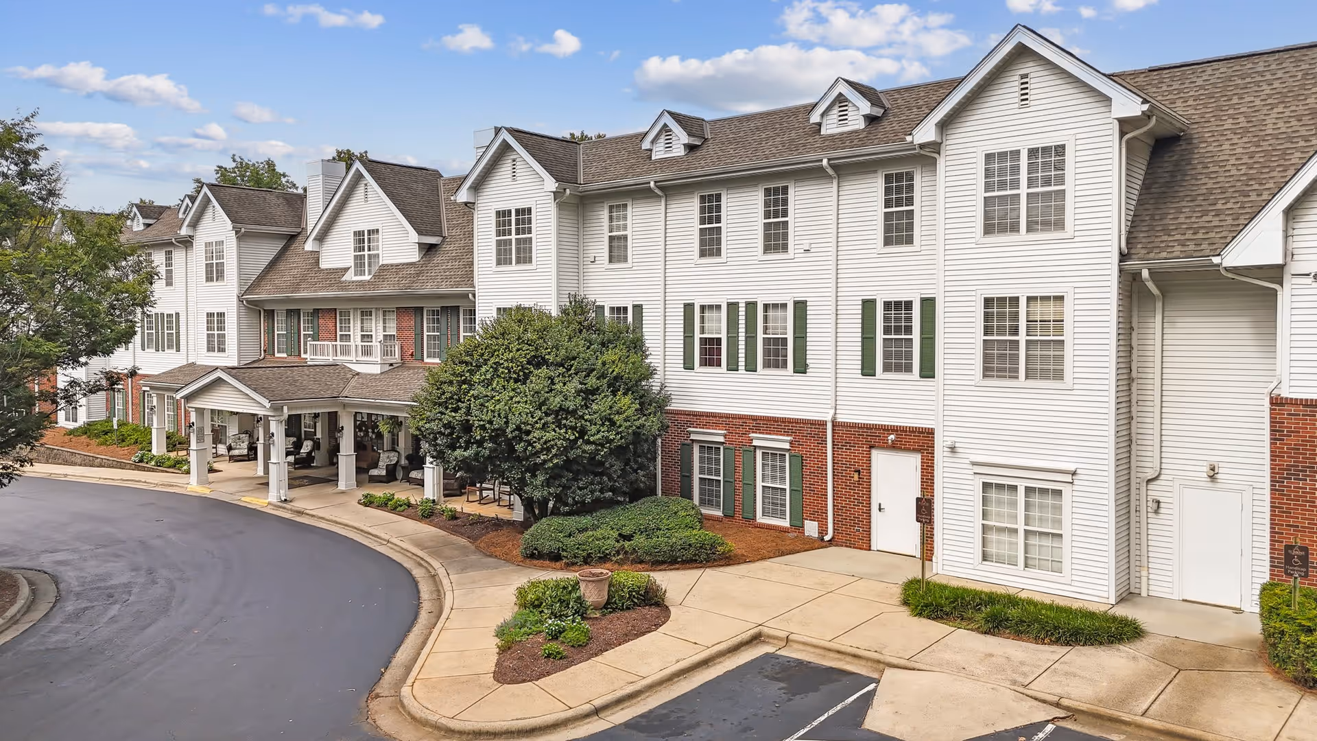 Exterior view of a multi-story senior living facility with white siding and brick accents, featuring multiple windows, a covered entrance with columns, landscaped bushes, and a curved driveway under a partly cloudy sky.