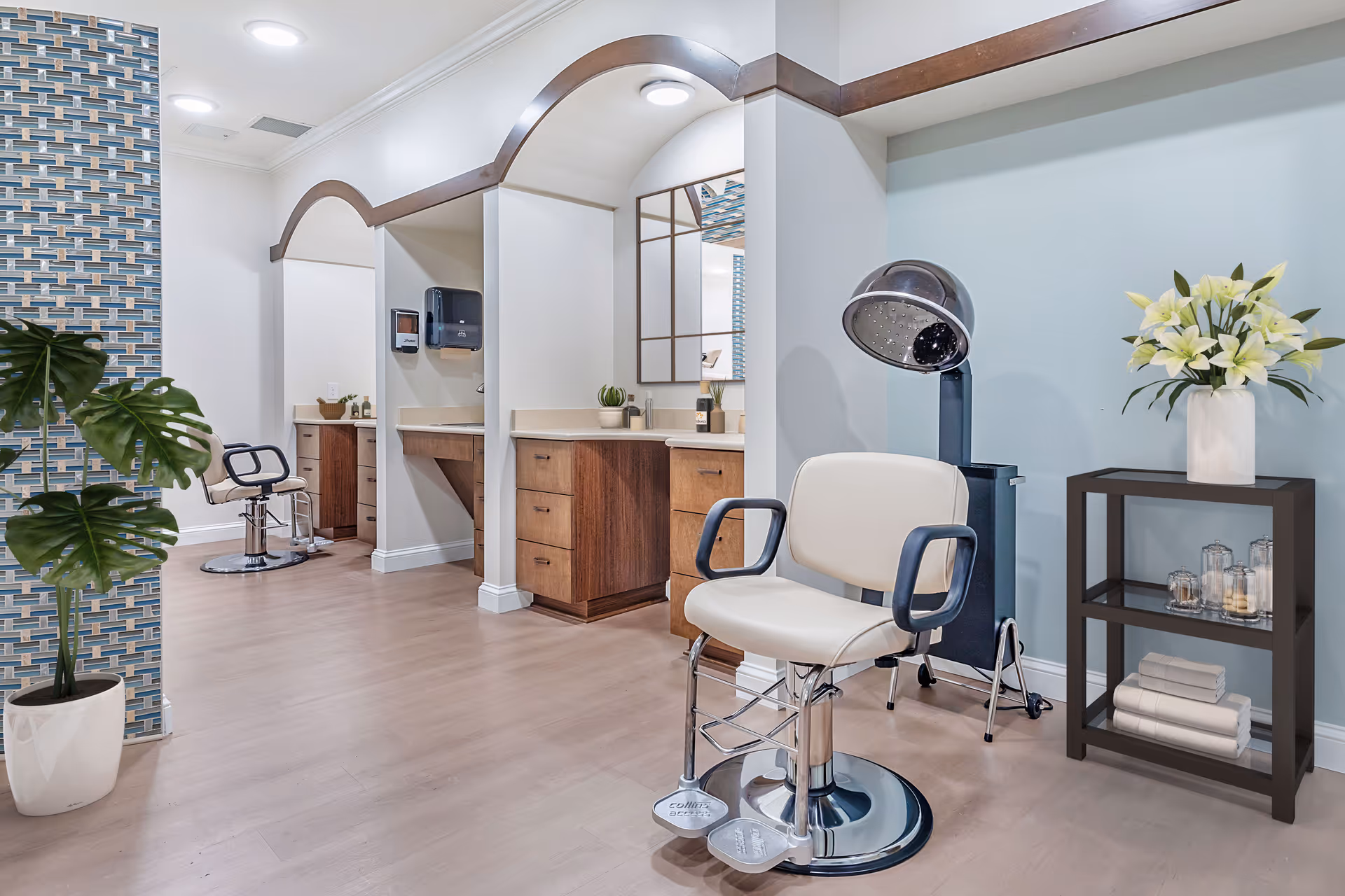 Bright senior living salon area with styling chairs, a hooded hair dryer, mirrors, plants, and shelving.