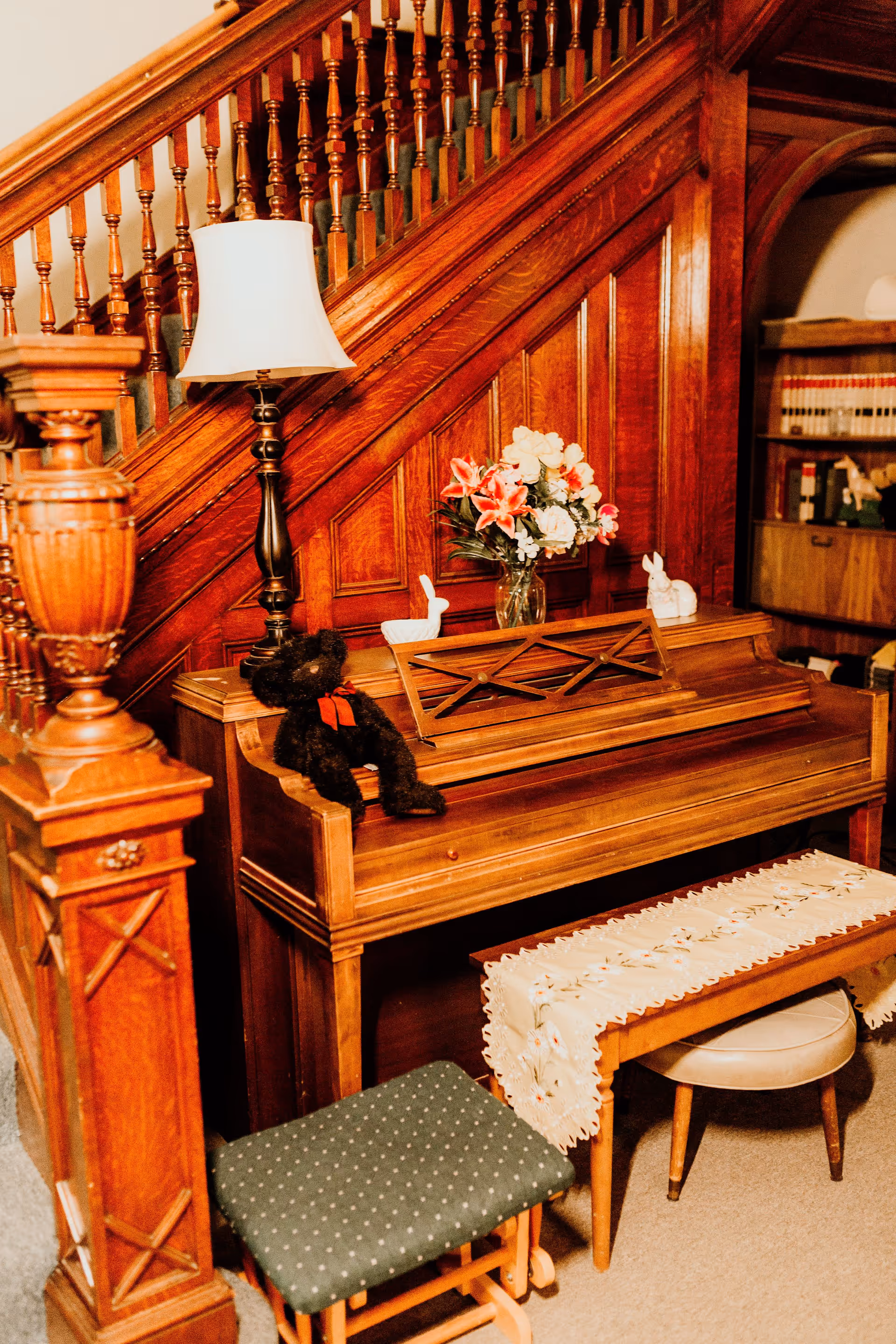 A wooden piano placed under a wooden staircase with a white lampshade on a dark lamp, a vase of flowers, a black teddy bear with a red bow, and two white ceramic rabbits on top. In front of the piano is a wooden bench covered with a decorative cloth and a green cushioned stool. A bookshelf with books is visible in the background.