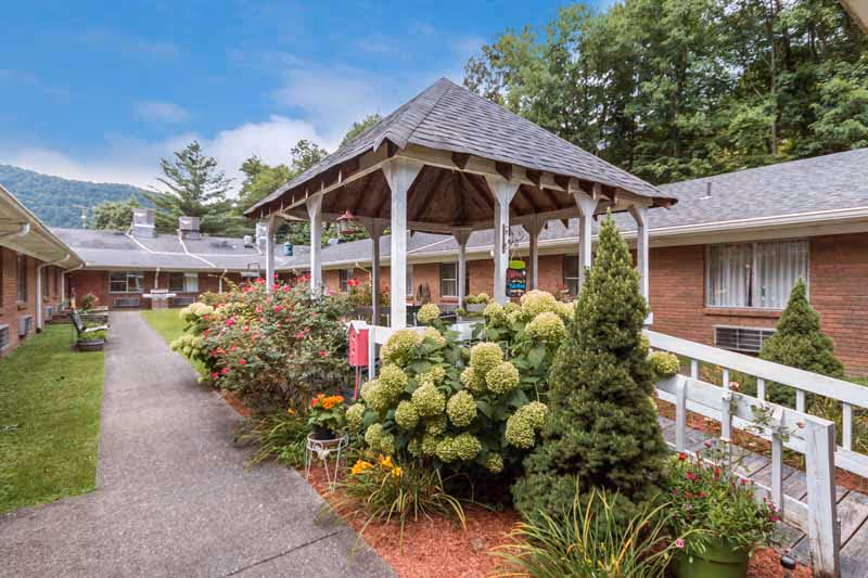 Outdoor courtyard area at Glasgow Hills featuring a wooden gazebo surrounded by lush green shrubs, flowering plants, and small trees. The courtyard is bordered by single-story brick buildings with windows and air conditioning units. A concrete walkway runs through the courtyard, and wooded hills are visible in the background under a partly cloudy sky.