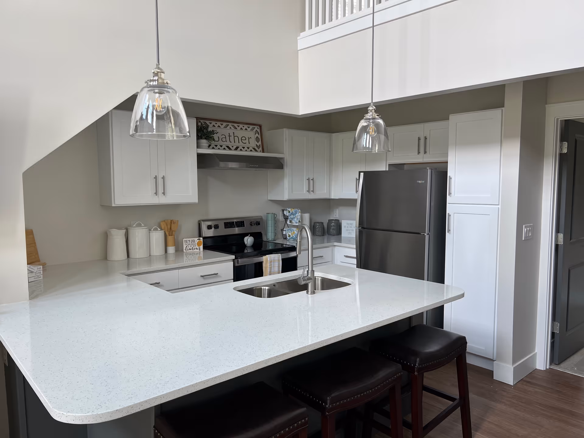 Bright modern kitchen with white cabinets, a large quartz island with sink and three bar stools, stainless steel appliances, and pendant lights.