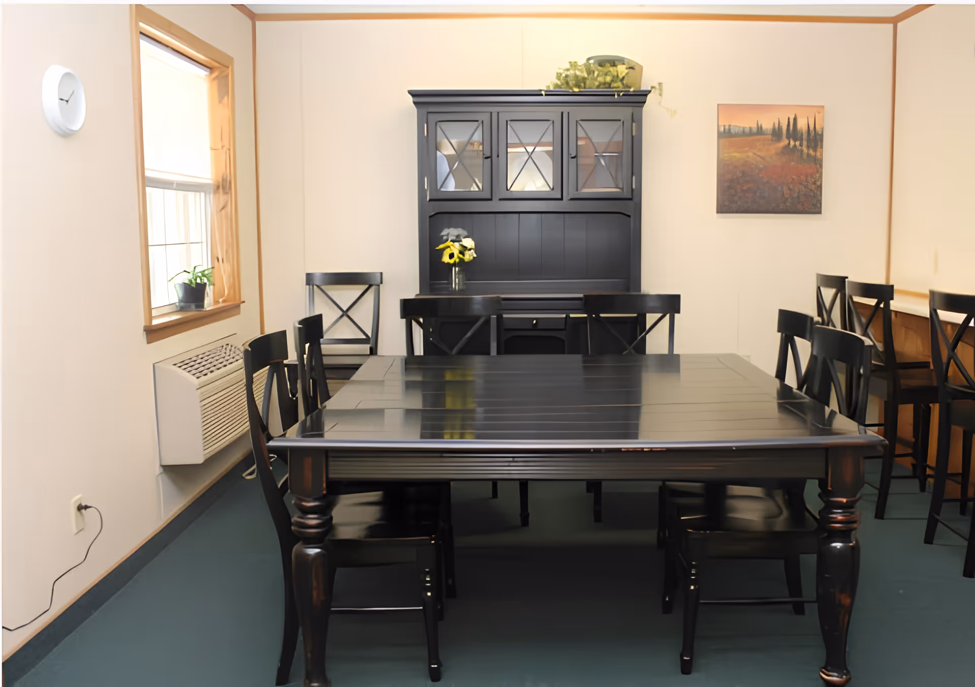 A dining room with a large dark wooden table surrounded by matching chairs. There is a black hutch cabinet against the back wall with glass-paneled doors and a vase with yellow flowers on the table. A window with a small potted plant and an air conditioning unit is on the left wall. A landscape painting hangs on the right wall.