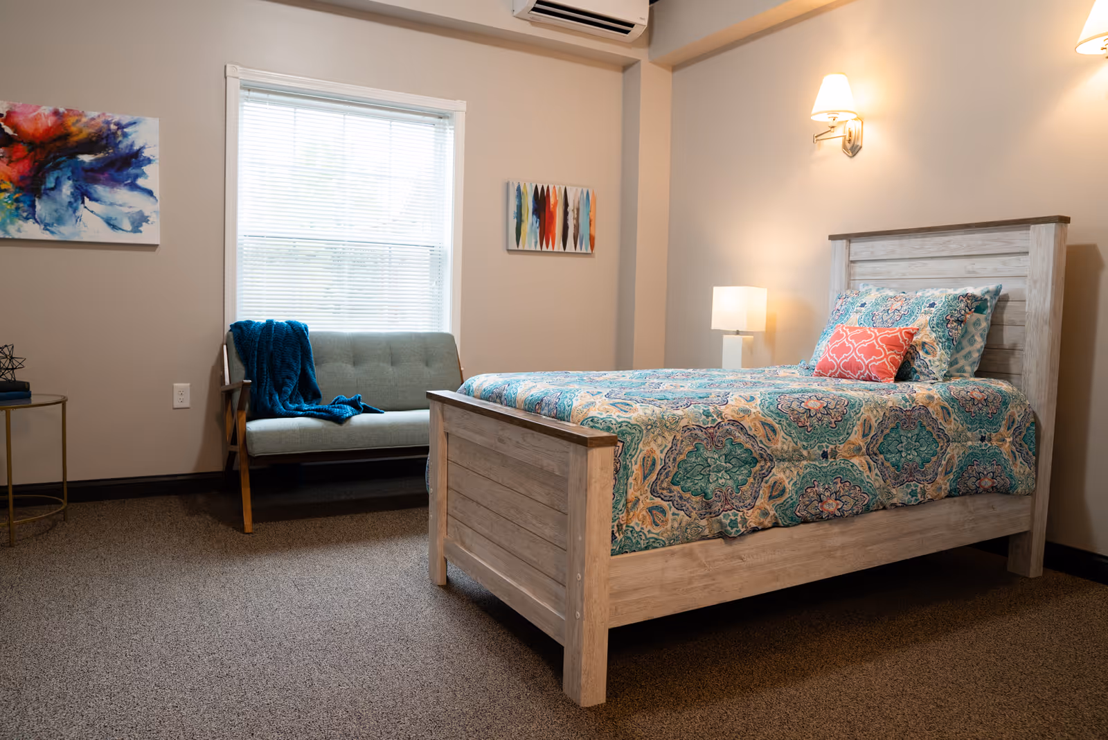 Well-lit bedroom with a wooden bed dressed in patterned bedding, a small loveseat by a window, and wall-mounted lamps.
