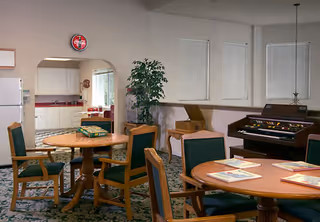 Interior view of a senior living facility common area with round wooden tables and green cushioned chairs. In the background, there is a kitchen area visible through an arched doorway, a large potted plant, a vintage record player, and an electric organ against the wall. The room has carpeted flooring and several windows with closed blinds.