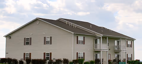 Two-story beige apartment building with balconies, shutters, and a gabled roof under a partly cloudy sky.