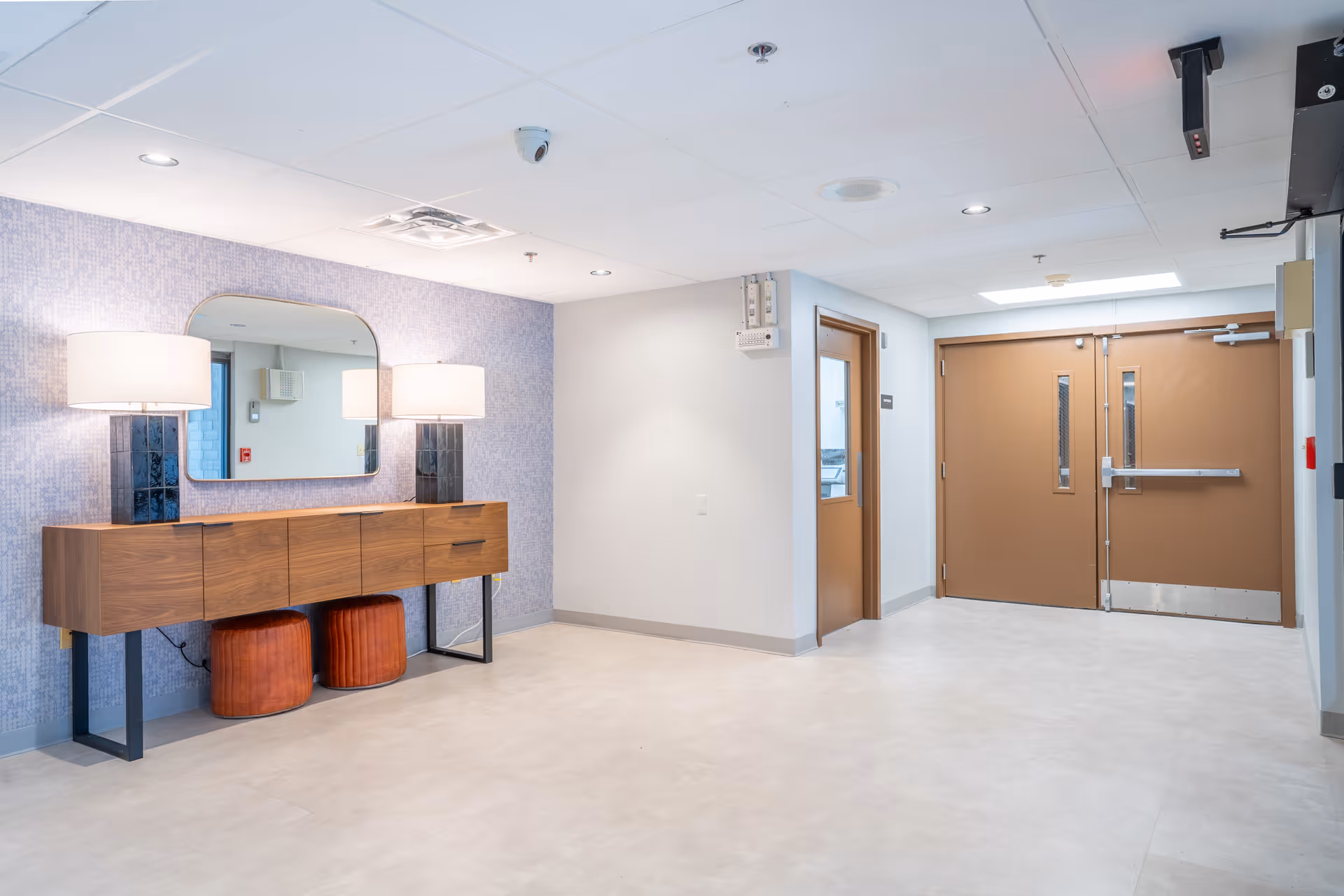 Bright nursing facility corridor with a wooden console table, two lamps and a mirror on the left and double brown doors at the far end.