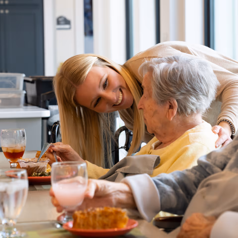 A smiling caregiver leans over and talks with an elderly woman as they sit at a dining table with plates and drinks.