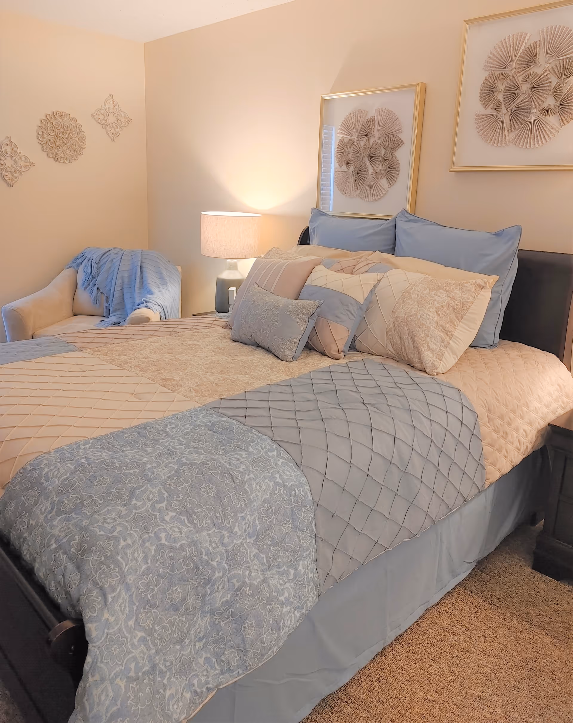 A neatly made bedroom with pastel blue and beige bedding, multiple pillows, a bedside lamp, an upholstered chair with a throw, and framed wall art.