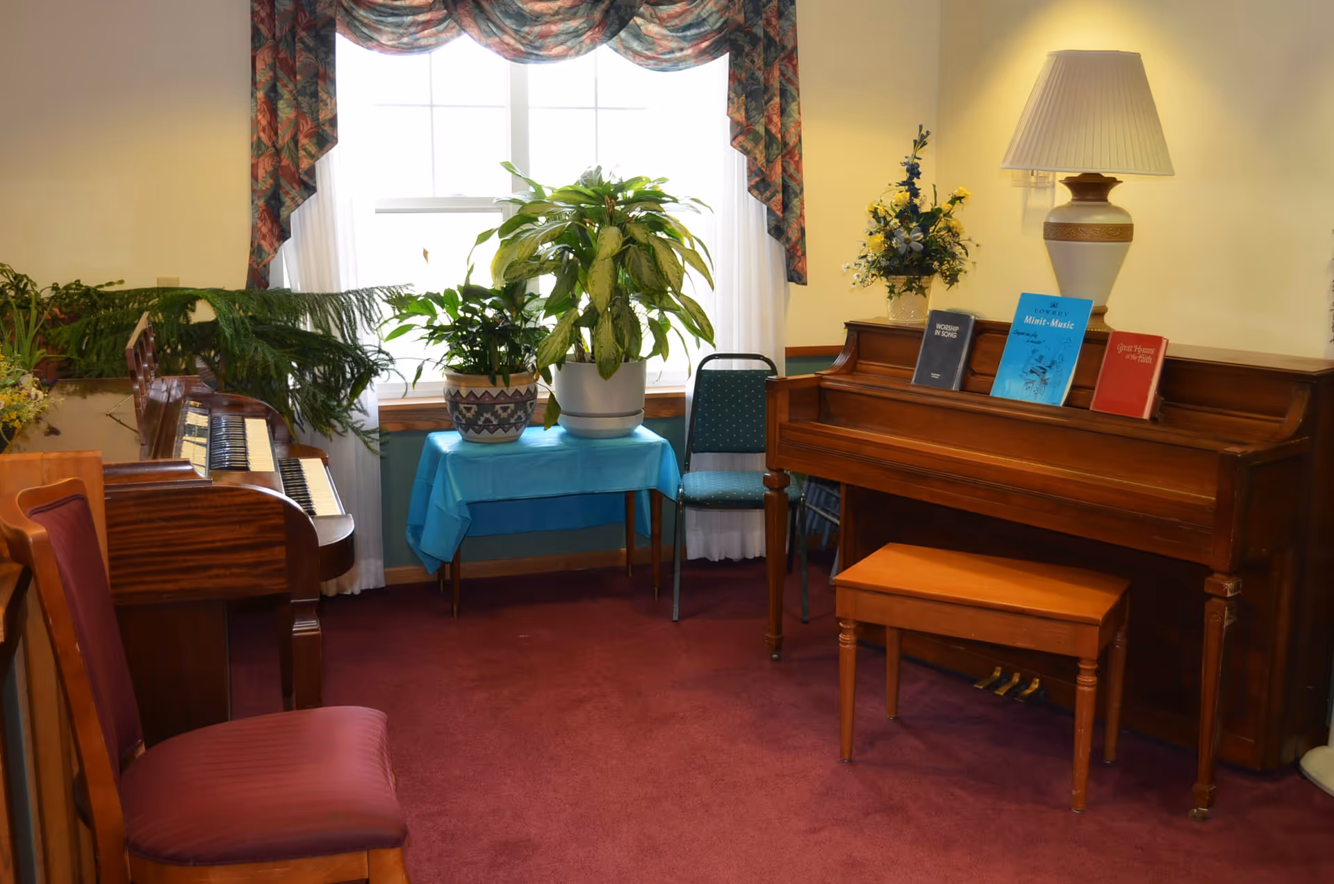 A cozy interior music room with two pianos, sheet music, chairs, potted plants and a lamp by a window.