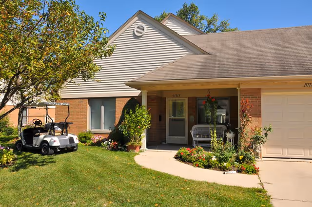 Exterior view of a single-story residential building with a brick and siding facade, a small front porch with a white bench, a garden with flowers and shrubs, a driveway, and a golf cart parked on the lawn under a tree.