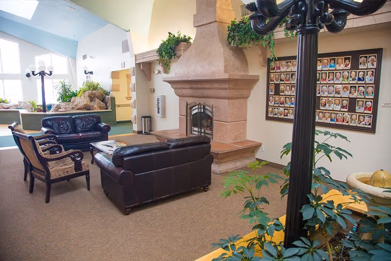 A cozy living room area with a large stone fireplace, two black leather sofas, and two upholstered armchairs. The room has beige carpeting, several green plants, and a wall display featuring many framed photographs of people. Large windows in the background let in natural light, and there is a rock water feature near the windows.