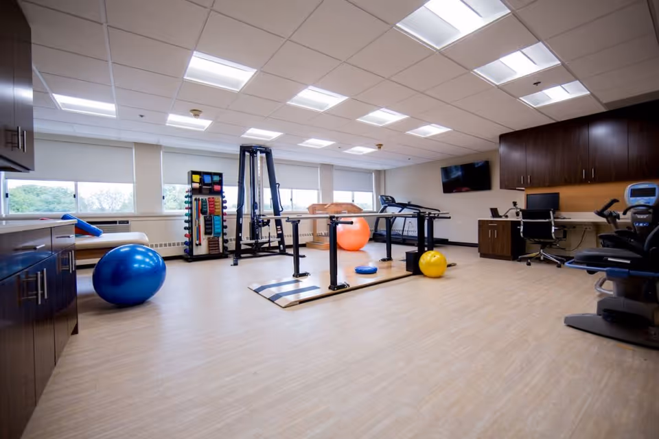 A spacious physical therapy or exercise room with large windows, exercise balls in blue, orange, and yellow, parallel bars for walking support, a rack with various exercise equipment, a treadmill, and a desk with a computer and chair. The room has light wood flooring, white ceiling tiles with fluorescent lights, and dark wood cabinetry along the walls.