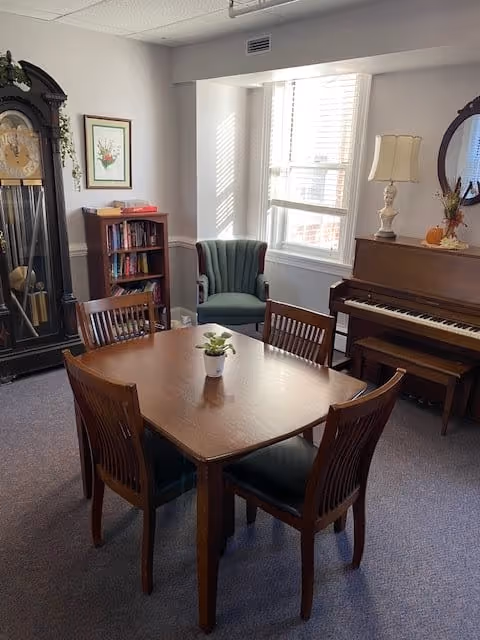 A cozy room with a wooden table and four chairs in the center, a green upholstered armchair by a window with white blinds, a wooden piano with a lamp and decorative items on top, a tall grandfather clock, and a small bookshelf with books and framed artwork on the wall.