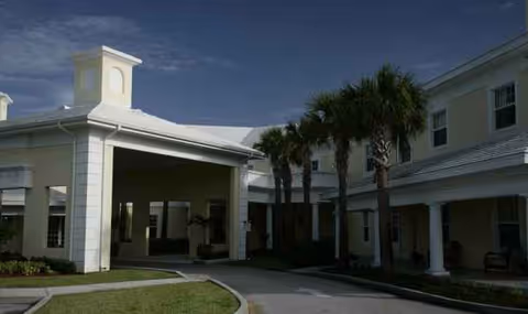 Exterior view of a two-story assisted living facility with a covered entry and palm trees.