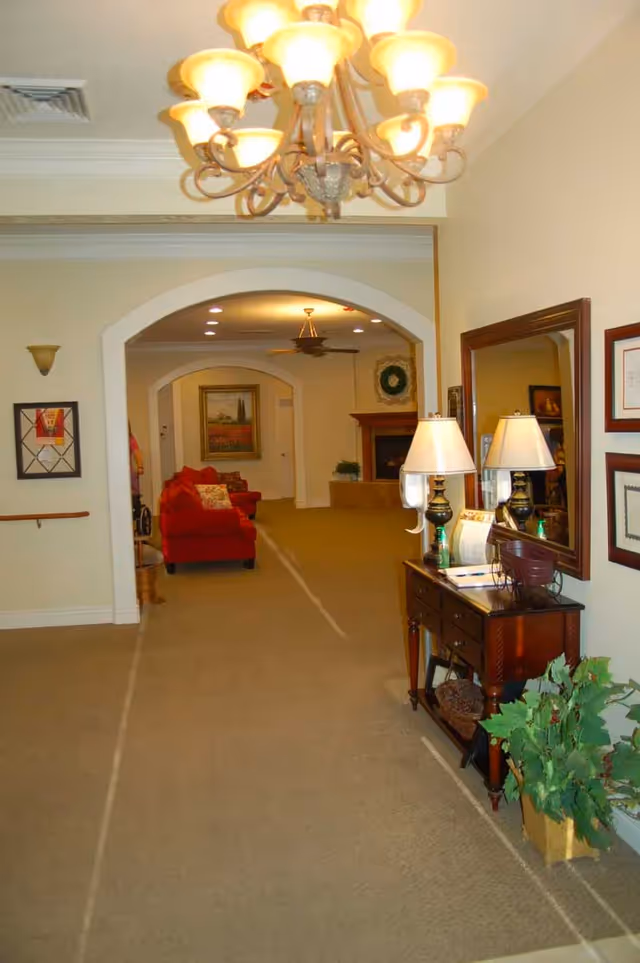 Interior view of a senior living facility hallway with beige carpet and walls, featuring a wooden console table with two lamps and decorative items on the right side. There is a large mirror above the table and framed pictures on the wall. In the background, there is a red armchair, a fireplace with a wreath above it, and a ceiling fan. A chandelier with multiple lights hangs from the ceiling near the foreground.