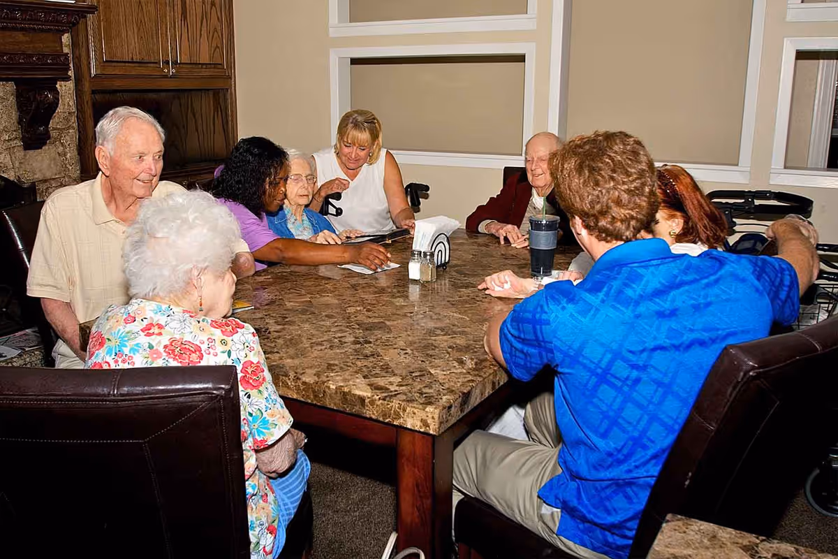 A group of elderly residents and caregivers seated around a large marble-topped table in a communal dining/activity room.