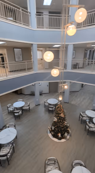 Interior view of a spacious two-story common area in a senior living facility with round tables and chairs arranged on the ground floor. A decorated Christmas tree is placed in the center. Modern spherical pendant lights hang from the ceiling. The upper floor has railings and framed artwork on the walls.