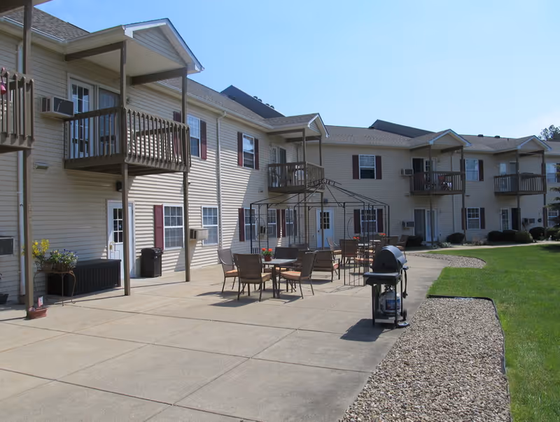 Outdoor patio area of a senior apartment complex with multiple tables and chairs arranged under a metal gazebo frame. There is a barbecue grill on the concrete patio, and the building has beige siding with balconies and windows. The area is bordered by a lawn and a strip of rocks.