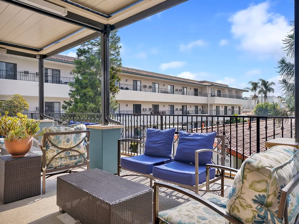 Outdoor balcony seating area with cushioned chairs and a small table, overlooking a multi-story building with balconies and a clear blue sky.