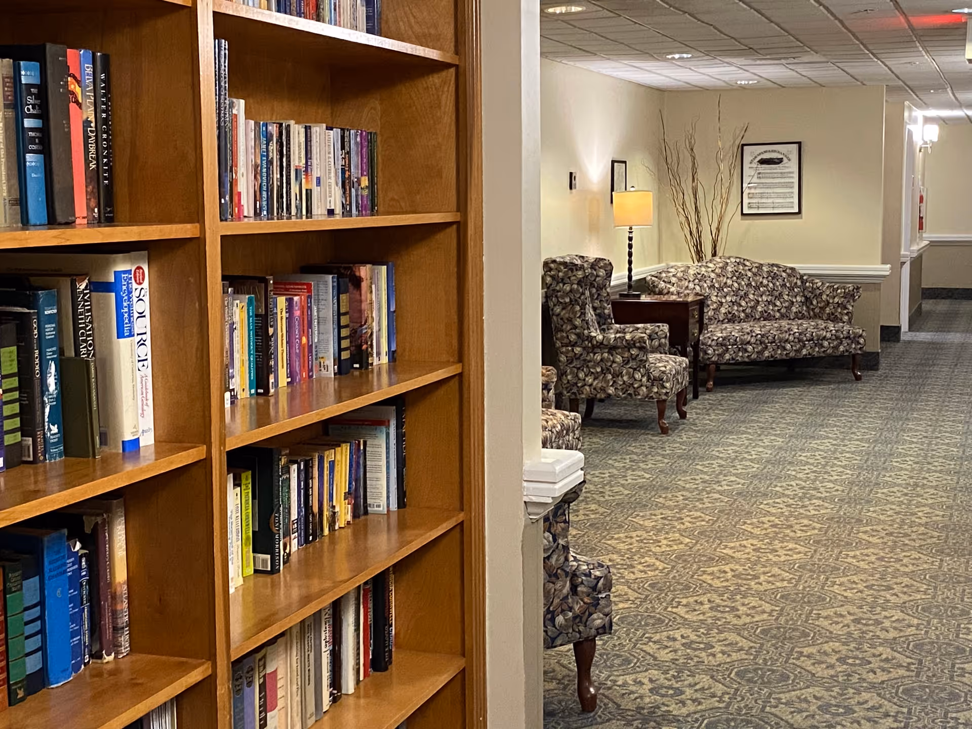 Interior view of a senior living facility hallway with a wooden bookshelf filled with books on the left and a seating area with patterned armchairs and a sofa, a side table with a lamp, and decorative branches on the right.