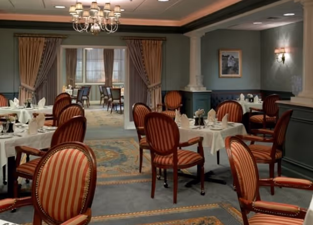 Elegant dining room with round tables covered in white tablecloths, set with napkins and tableware. The room features striped upholstered chairs, a chandelier, framed artwork on the walls, and large windows with curtains in the background.