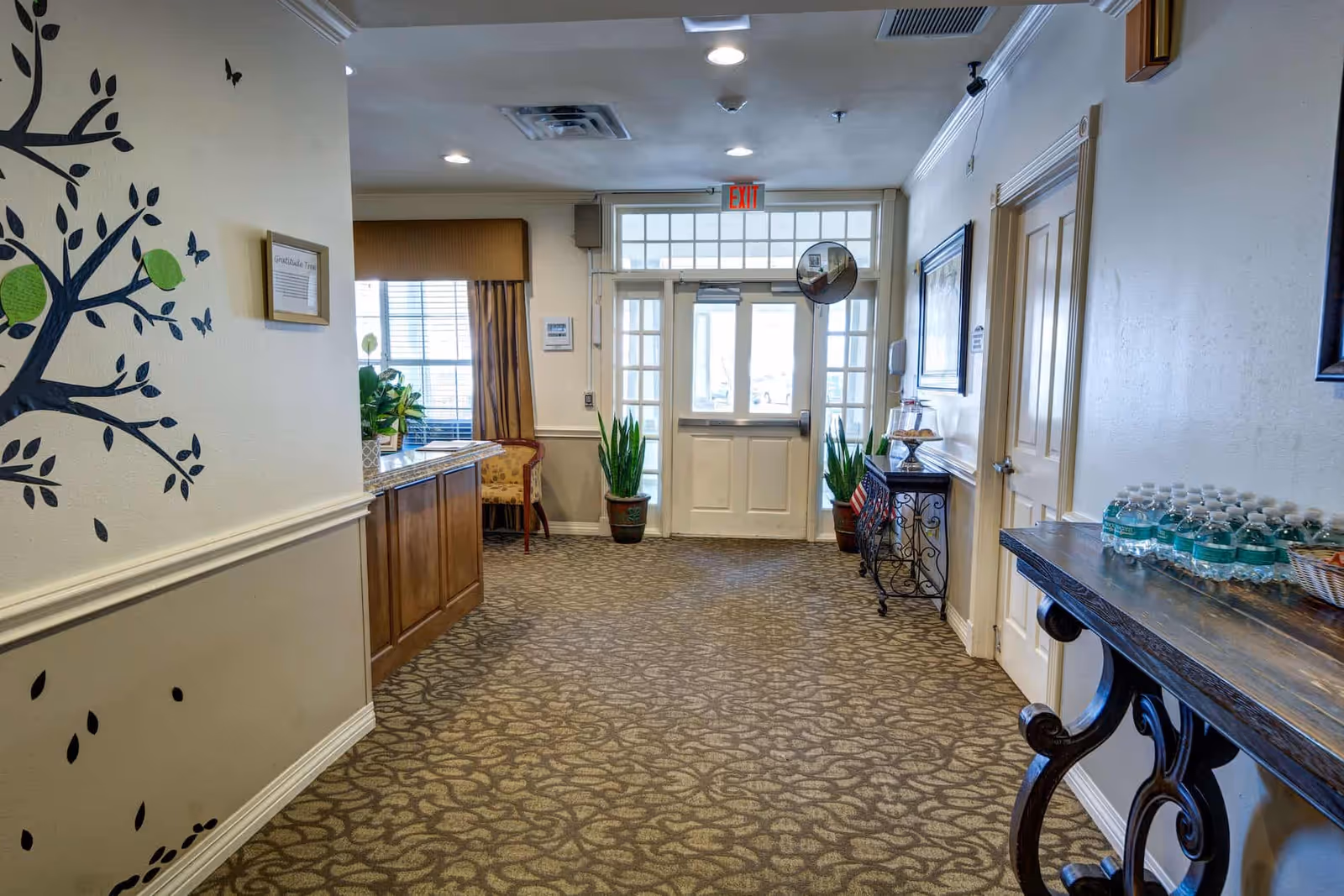 Interior lobby/hallway of a senior living facility with double entrance doors, decorative tables, plants, wall art, and bottled water on a side table.