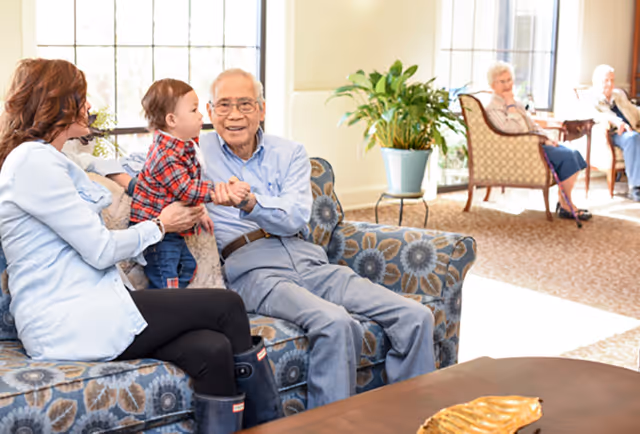 An elderly man sitting on a patterned couch holding hands with a young child who is standing on the couch, while a woman sits next to them. In the background, two elderly women are seated on chairs near large windows with sunlight streaming in. A potted plant is placed on a small stand near the wall.