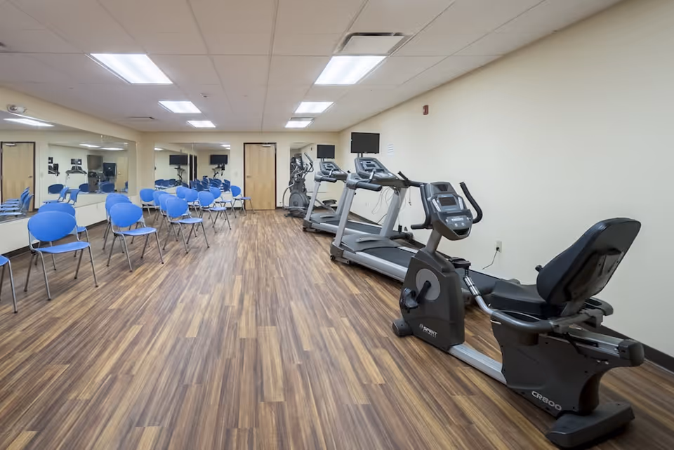 A fitness room with wooden flooring featuring exercise equipment including a recumbent bike, two treadmills, and an elliptical machine. On the left side of the room, there are several blue chairs arranged in rows facing the exercise machines. The walls are light-colored, and there are large mirrors on the left wall reflecting the room. The ceiling has recessed lighting panels.