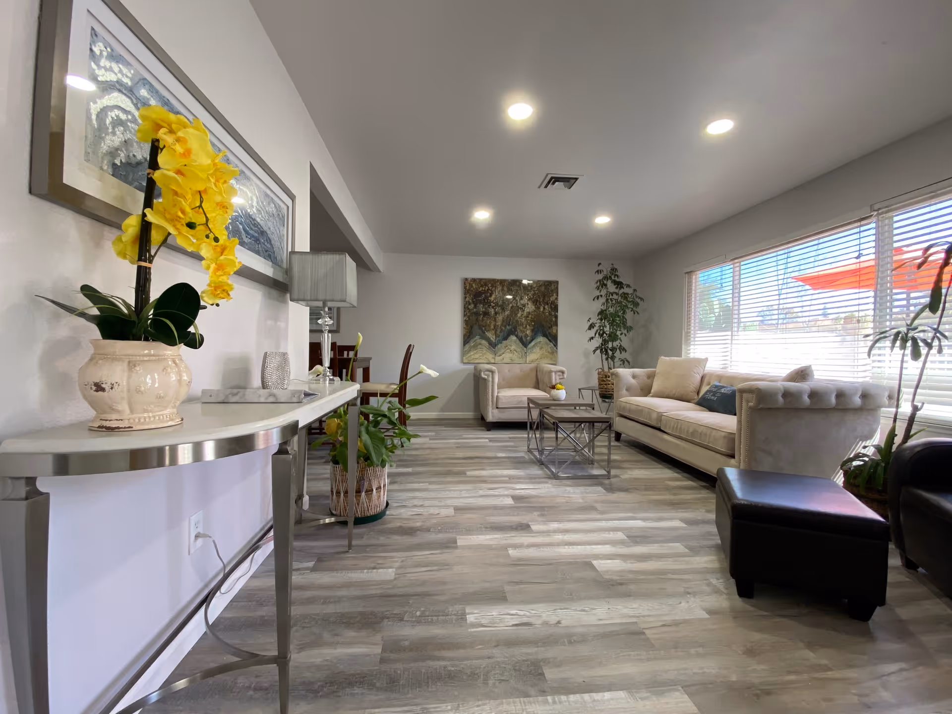 A bright and modern living room with light gray wood flooring, a beige tufted sofa, a matching armchair, a glass coffee table with a metal frame, and several potted plants. Large windows with blinds allow natural light to fill the room. A console table with a yellow orchid and decorative items is positioned along one wall, and a large abstract painting hangs on the far wall.