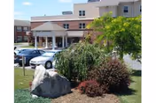 Exterior view of a multi-story retirement community building with a covered entrance, several parked cars, and landscaped greenery including bushes, a tree, and a large rock in the foreground.