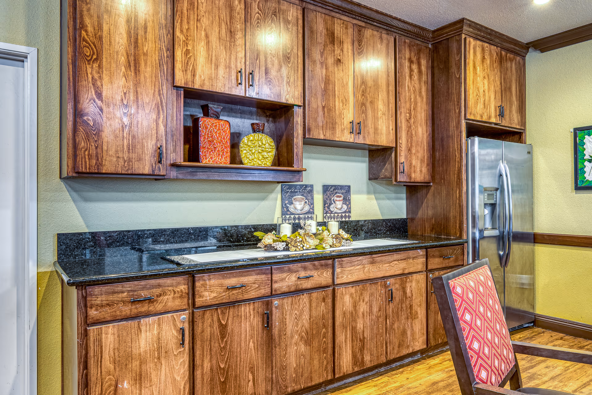 Interior view of a kitchen area with wooden cabinets, a black granite countertop, a stainless steel refrigerator, and a chair with a red patterned cushion. Decorative items including two vases and a floral arrangement with candles are placed on the countertop.