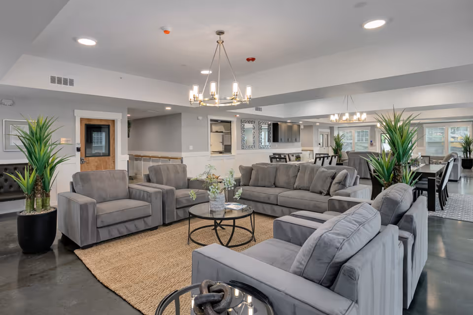 A spacious and modern senior living facility common area with multiple gray upholstered sofas and armchairs arranged around a round glass coffee table on a woven rug. The room features contemporary chandeliers, large potted plants, and a dining area with tables and chairs in the background. The walls are painted light gray with white trim, and the floor is polished concrete.