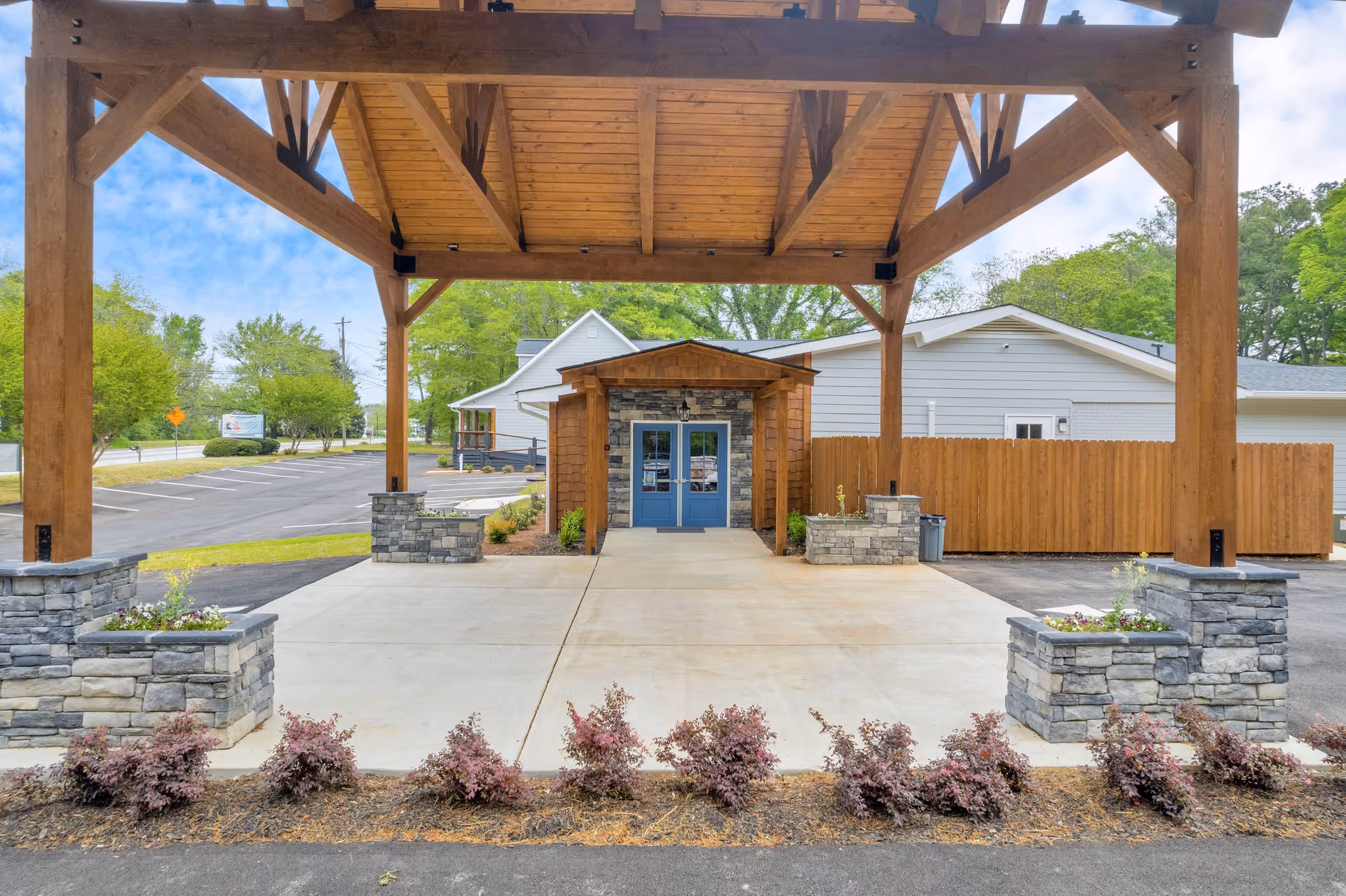Entrance of a senior living facility with a wooden pergola structure supported by stone pillars, a concrete driveway, landscaped bushes, and a building with blue double doors and stone accents under a partly cloudy sky.