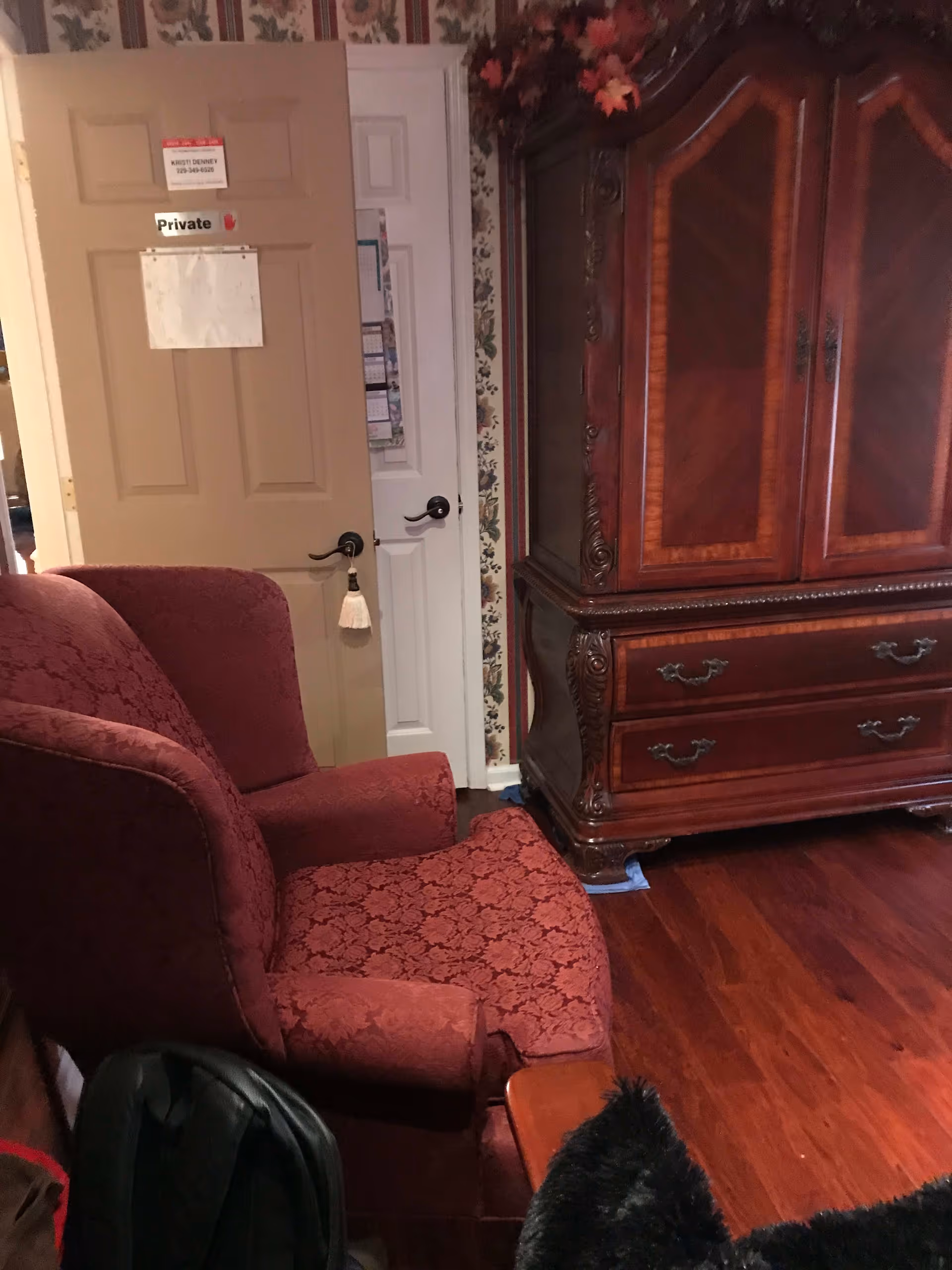 Interior room with a red patterned armchair, a wooden armoire with decorative carvings, floral wallpaper, and two closed doors, one labeled 'Private'. The floor is wooden and there are some items partially visible in the foreground.