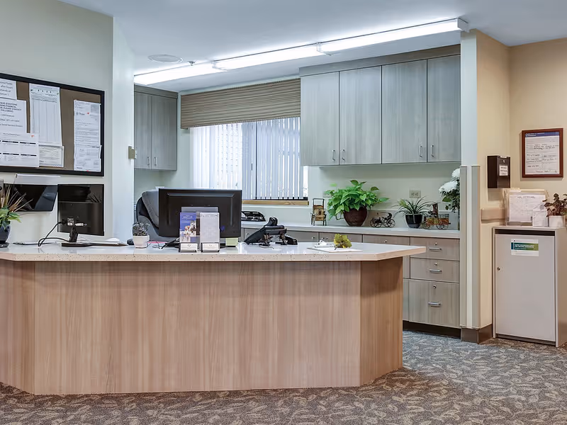Reception desk area in a senior living facility with a wooden counter, two computer monitors, office supplies, potted plants, and cabinets in the background. A window with vertical blinds is behind the desk, and bulletin boards with notices are on the walls.