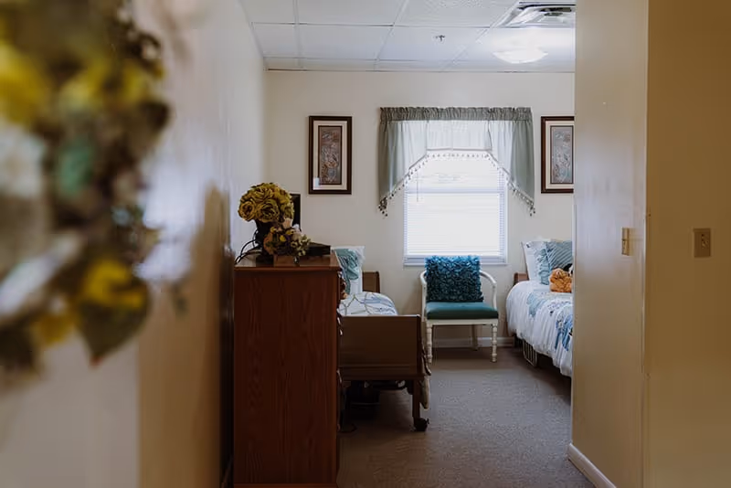 View down a hallway into a small bedroom with two beds, each with white and blue bedding. Between the beds is a green chair with a blue cushion. A window with a valance is centered on the back wall, flanked by two framed pictures. A wooden dresser with a flower arrangement is on the left side of the room.