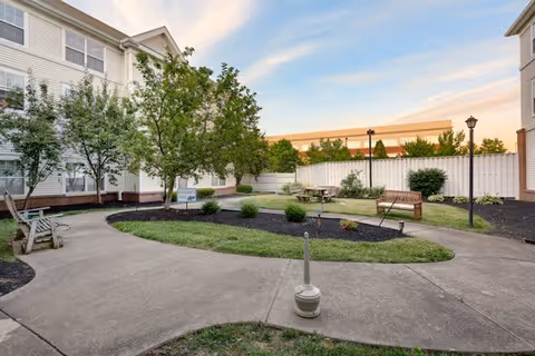 Outdoor courtyard area with paved walkways, benches, trees, and landscaped garden beds surrounded by multi-story residential buildings under a partly cloudy sky.