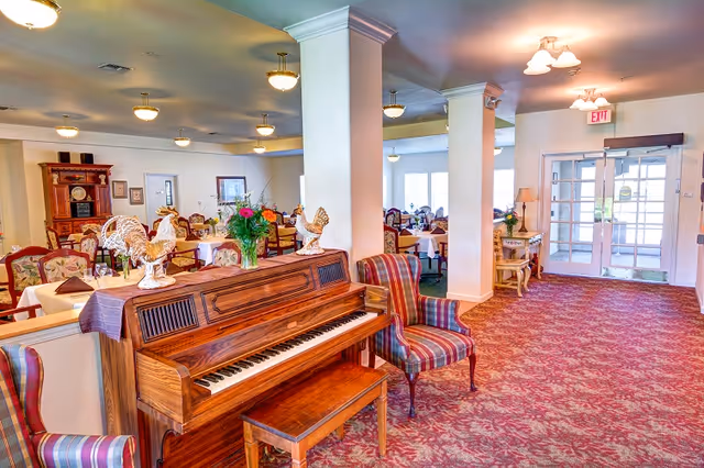 Interior view of a senior living facility dining area with multiple tables and chairs arranged for meals. In the foreground, there is a wooden upright piano with decorative rooster figurines and a vase of flowers on top. Next to the piano are two striped upholstered chairs. The room has carpeted floors with a red patterned design, white walls, and ceiling lights. Glass double doors are visible at the far end of the room.