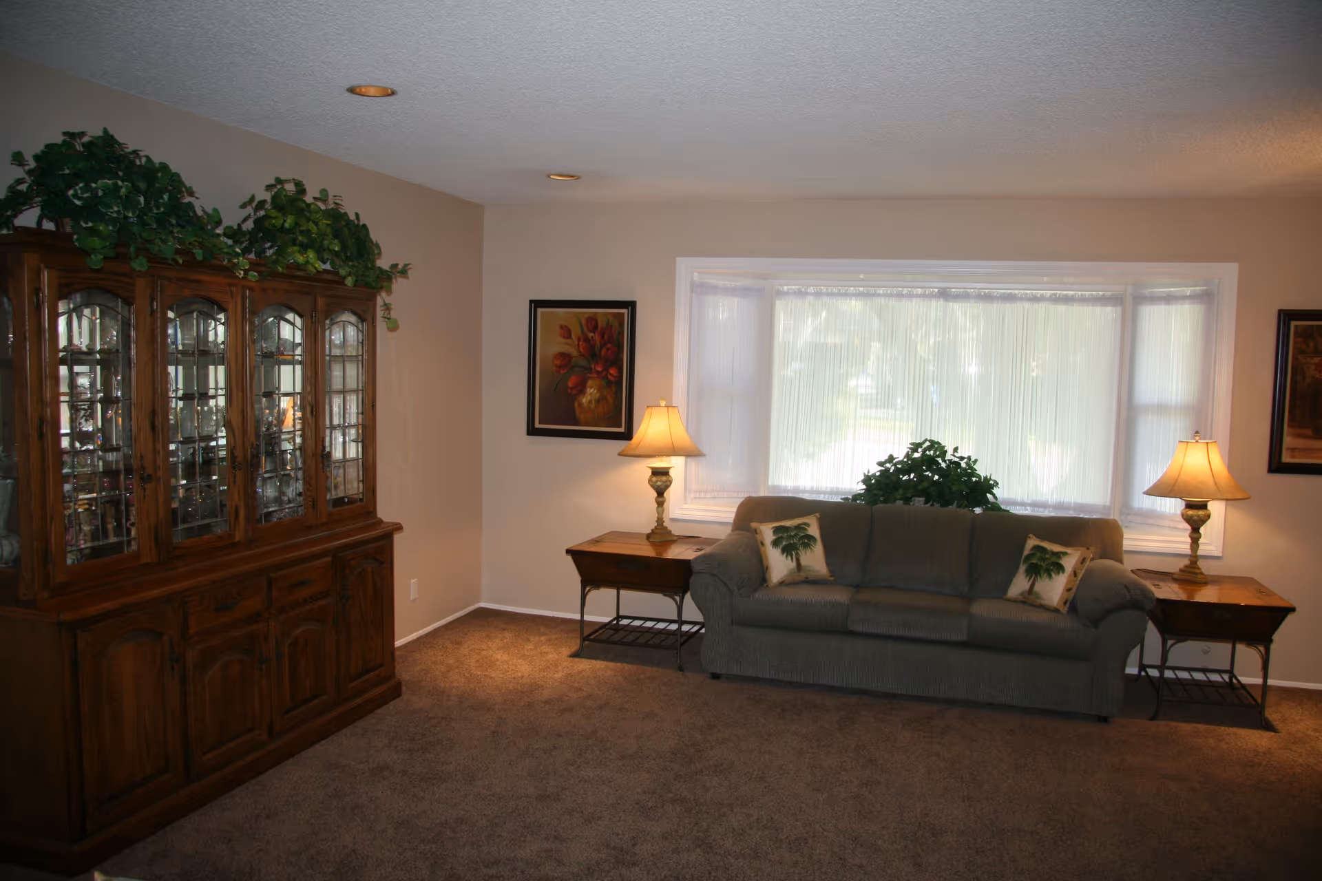 A living room with a sofa flanked by two table lamps beneath a large window and a wooden china cabinet on the left.
