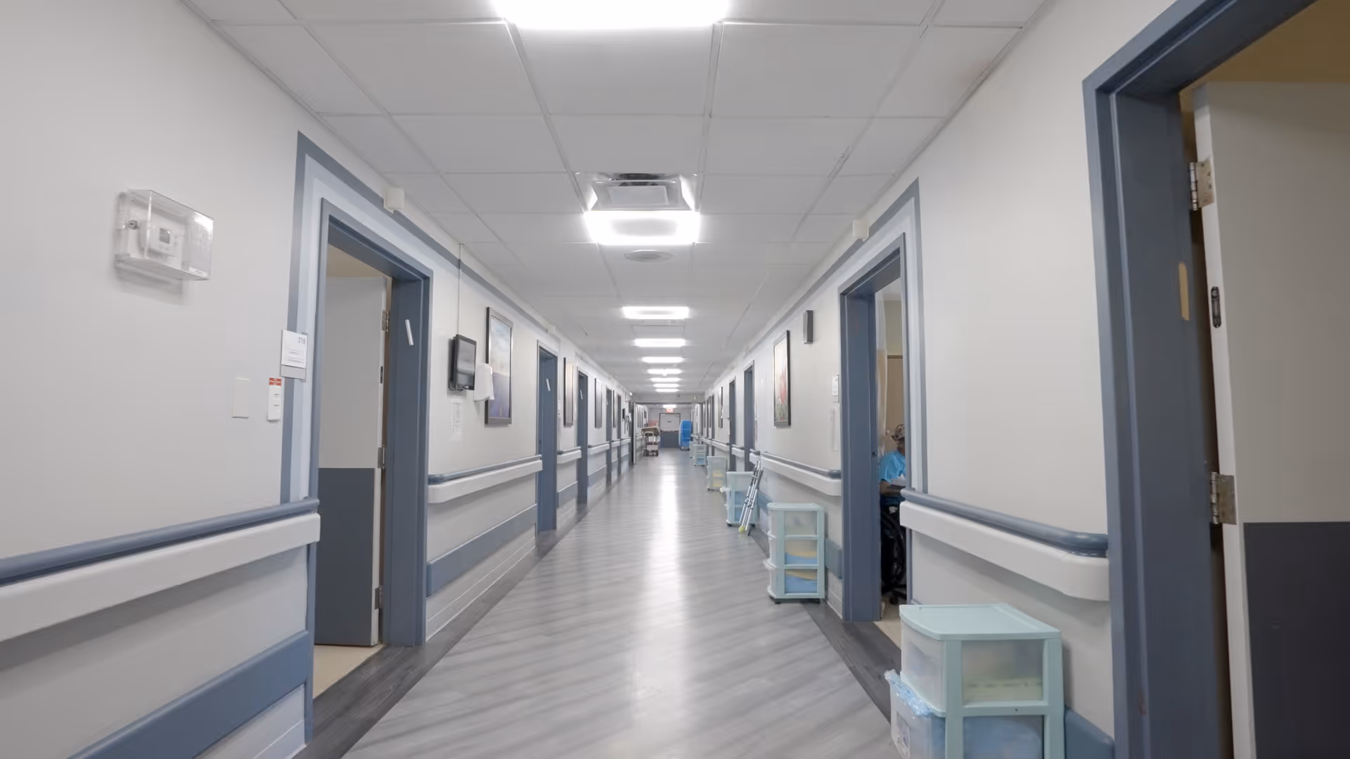 A long, clean hallway in a care facility with light gray walls and blue trim. Multiple open doorways line both sides of the corridor, with handrails running along the walls. The floor is covered with light gray wood-patterned vinyl. Several small storage carts are placed along the hallway, and fluorescent ceiling lights illuminate the space.