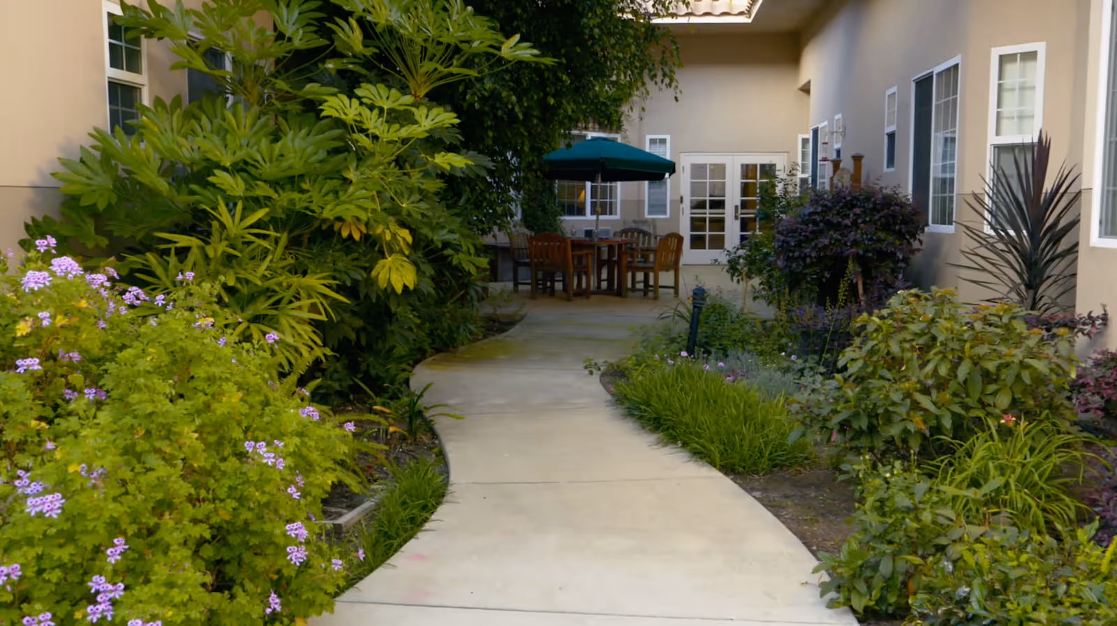 A paved walkway surrounded by lush green plants and flowers leading to an outdoor seating area with wooden tables and chairs under a green umbrella, adjacent to a beige building with multiple windows and a glass door.
