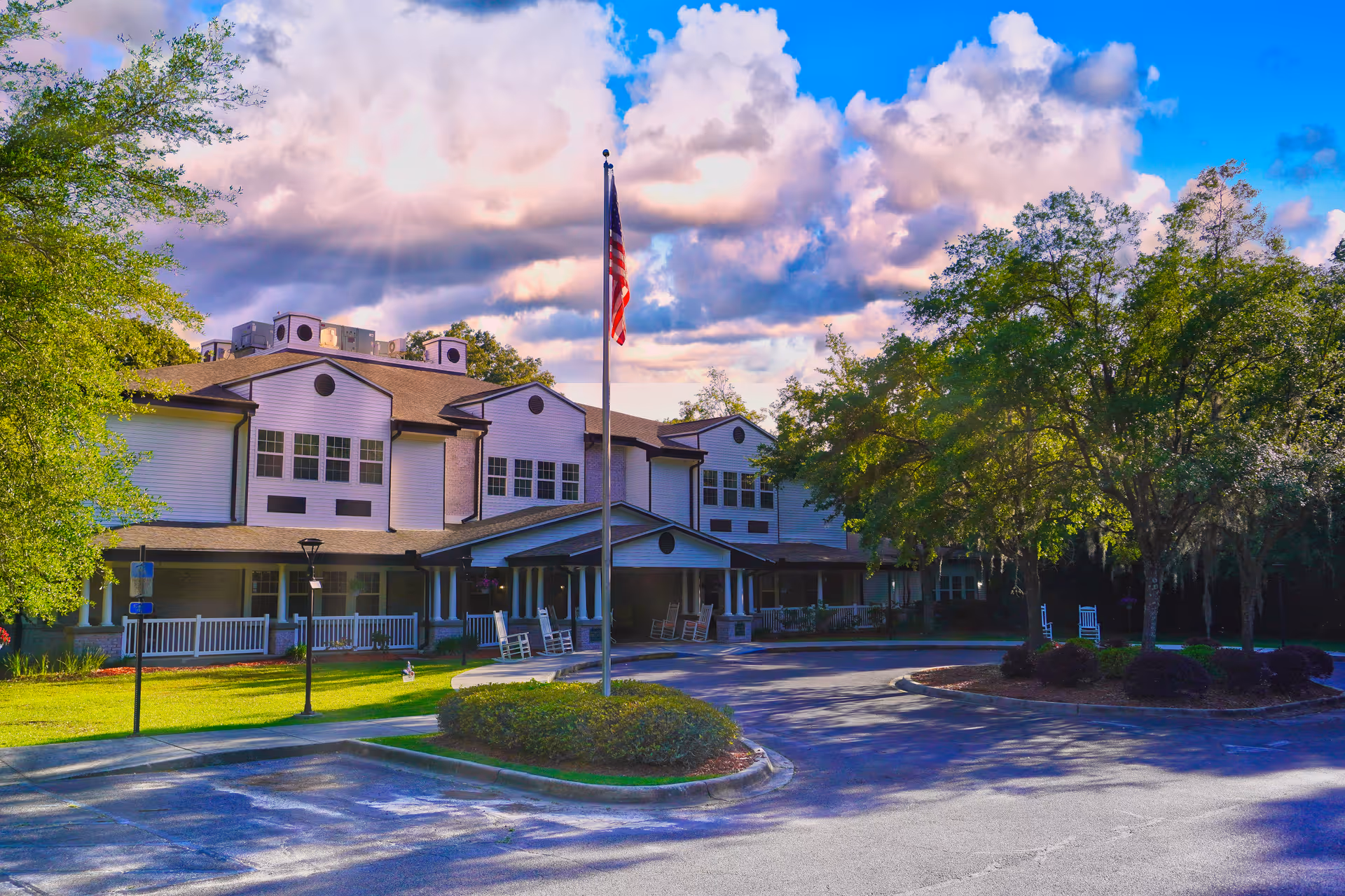 Exterior view of Sodalis Tallahassee Assisted Living facility showing a two-story building with white siding and multiple windows. There is a covered entrance with rocking chairs on the porch, a flagpole with an American flag in front, and trees and bushes surrounding the parking area under a partly cloudy sky.