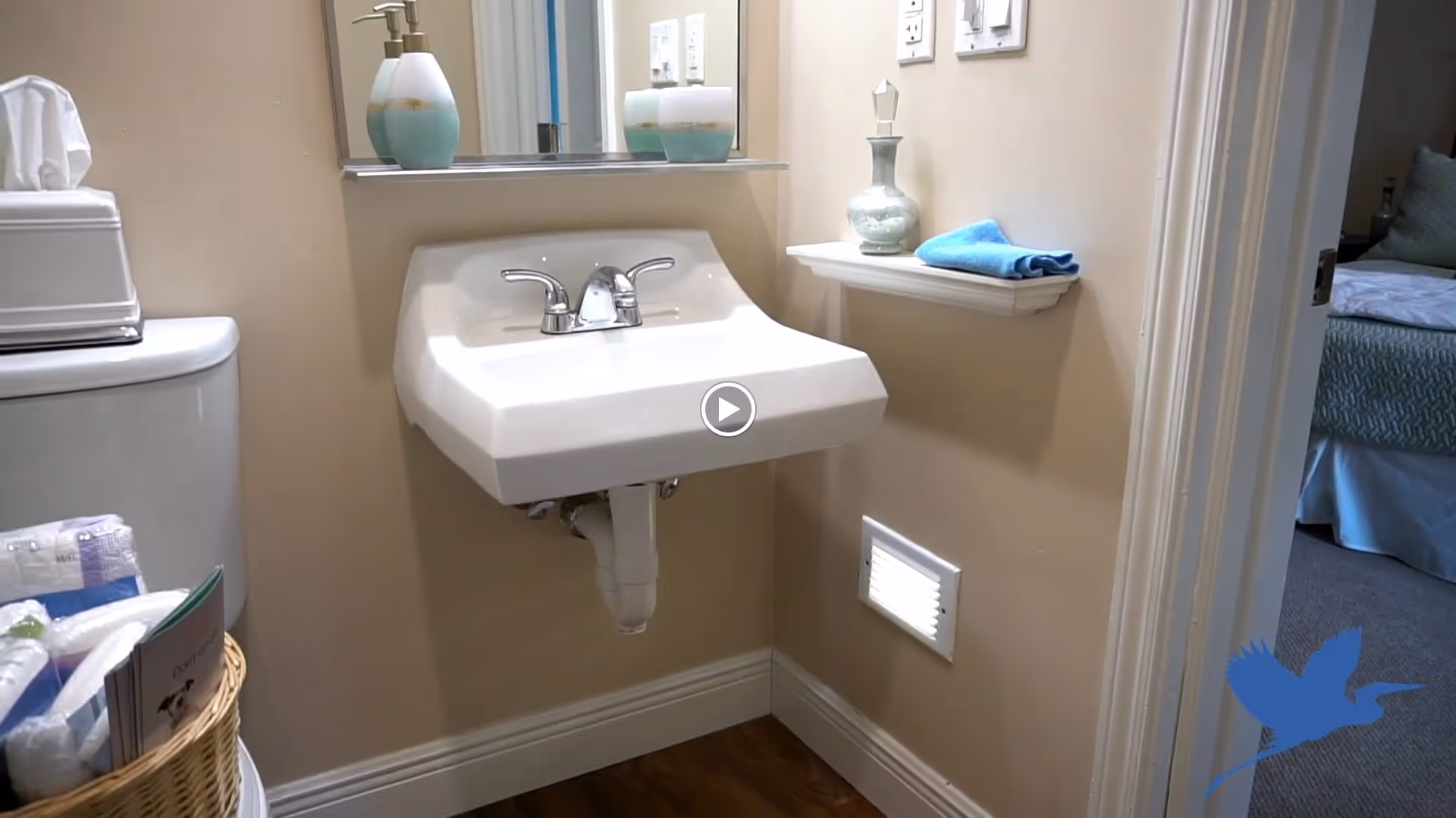 A small bathroom corner with a white wall-mounted sink, a mirror above it, a soap dispenser on the sink, and a small shelf holding a decorative bottle and a folded blue towel. To the left, part of a toilet with a tissue box on top and a basket with supplies is visible. The bathroom has beige walls and wooden flooring. A doorway on the right leads to a bedroom with a bed and pillows.