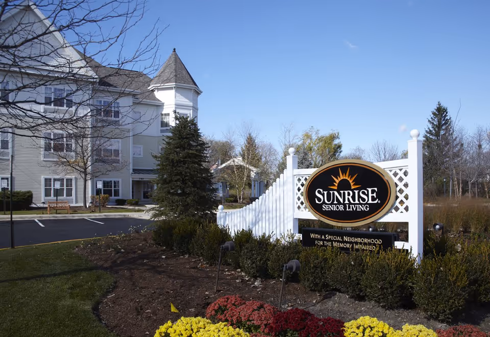 Exterior view of Sunrise Senior Living facility with a white multi-story building, a landscaped garden with colorful flowers, and a prominent sign reading 'Sunrise Senior Living' with a note about a special neighborhood for the memory impaired.